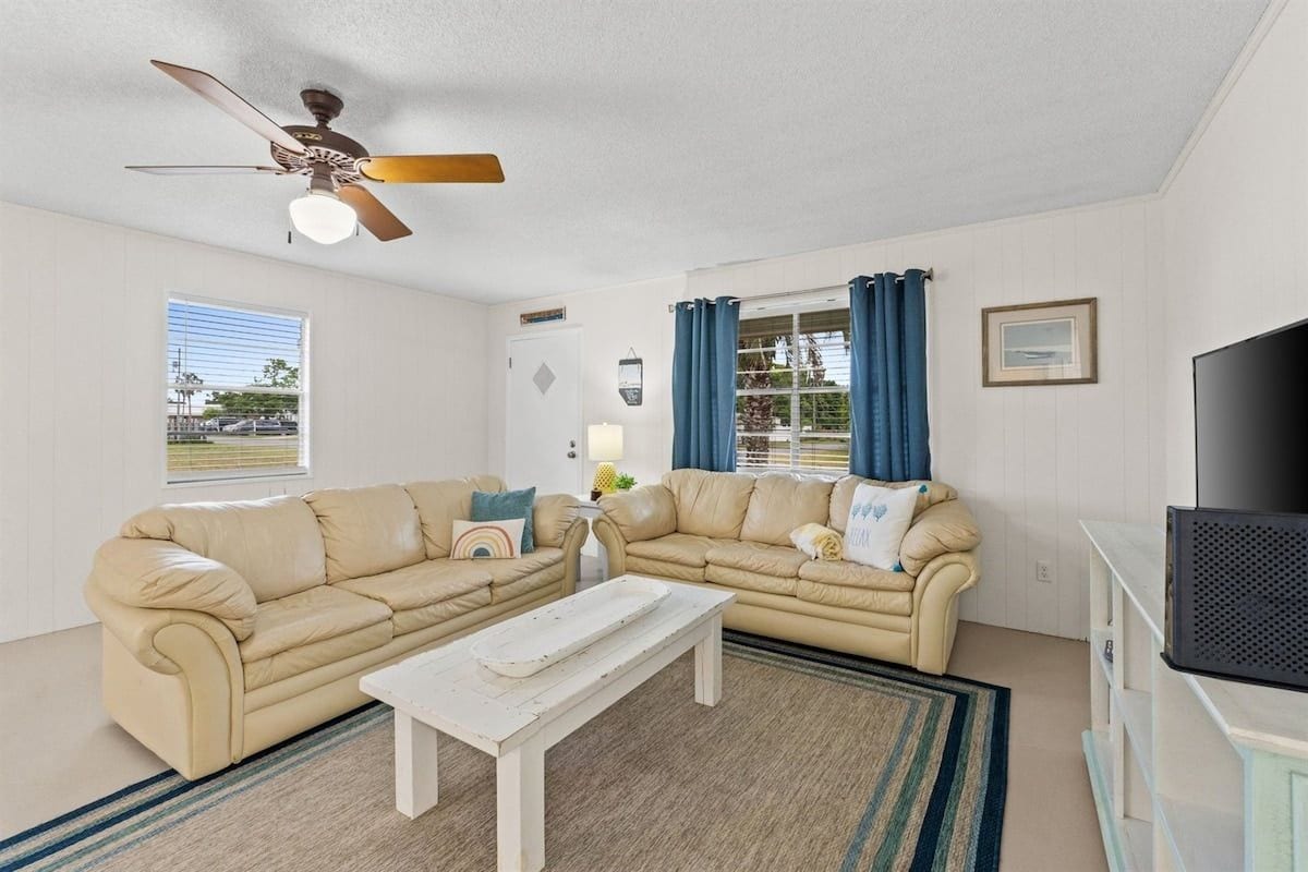 A living room with two beige sofas, a white coffee table, a ceiling fan, a TV, and a window with curtains.