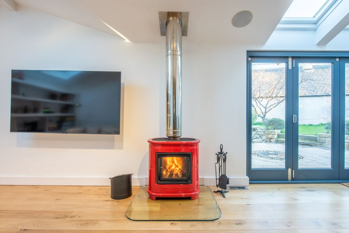 Un salon moderne avec un poêle à bois rouge, une table basse en verre et une grande fenêtre donnant sur l’extérieur.