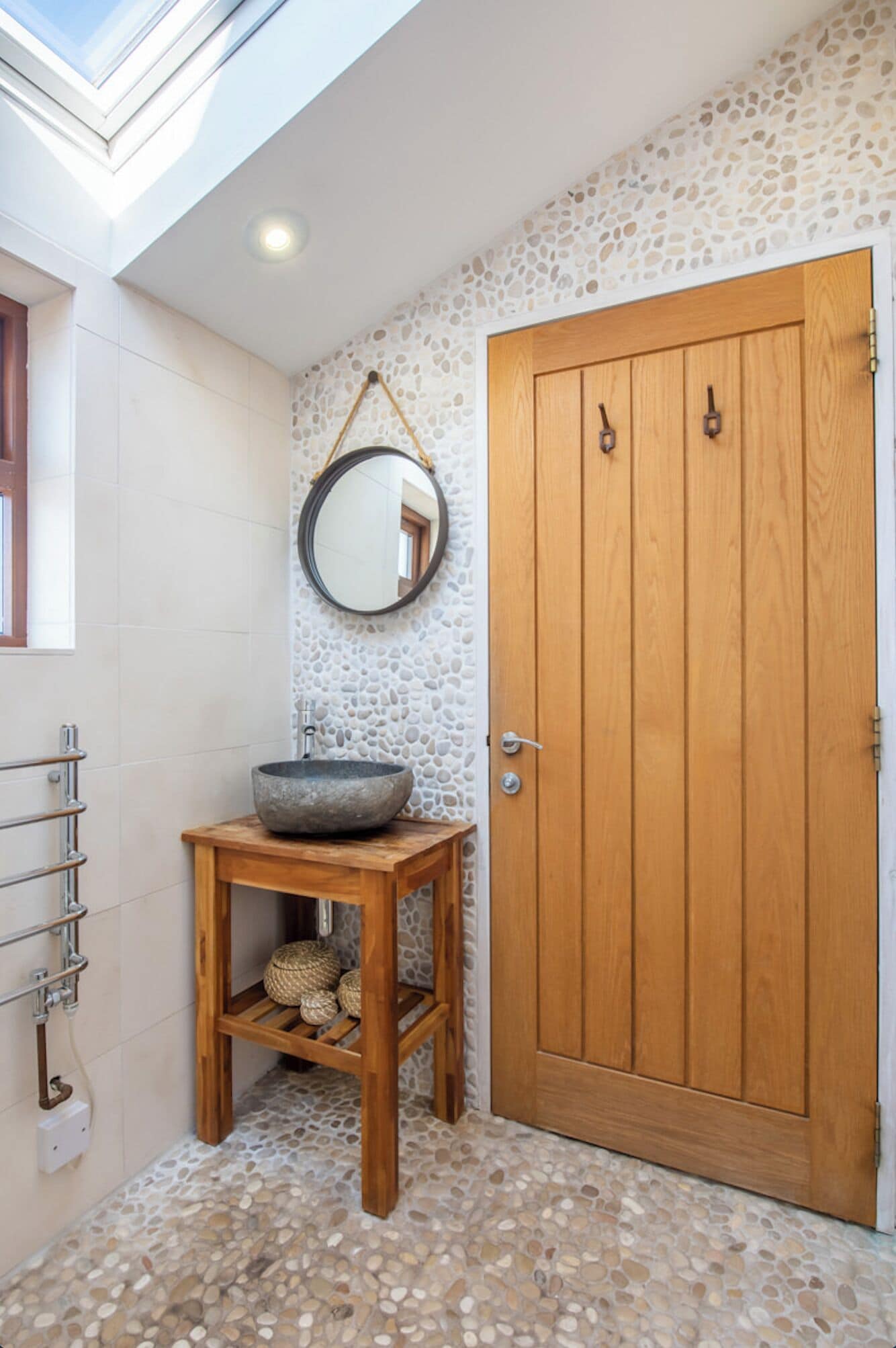 A bathroom with a stone mosaic floor, a wooden door, a round mirror, and a wooden sink with a bowl.