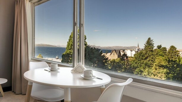 Una habitación con una mesa redonda y dos sillas, una ventana con vistas al mar y las montañas, y una cortina en el lado izquierdo.