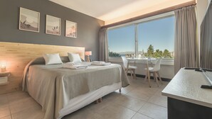 Habitación de hotel con cama, escritorio, silla, televisor y ventana con vista a árboles y edificios.