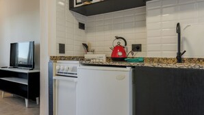 Una cocina compacta con paredes de azulejos blancos, encimera negra, refrigerador blanco, hervidor rojo y grifo negro.