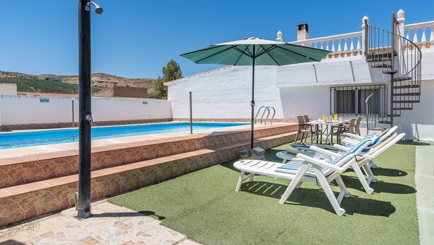 Zona de piscina amb un banyera d'hidromassatge, gandules i una taula amb ombrelles.
