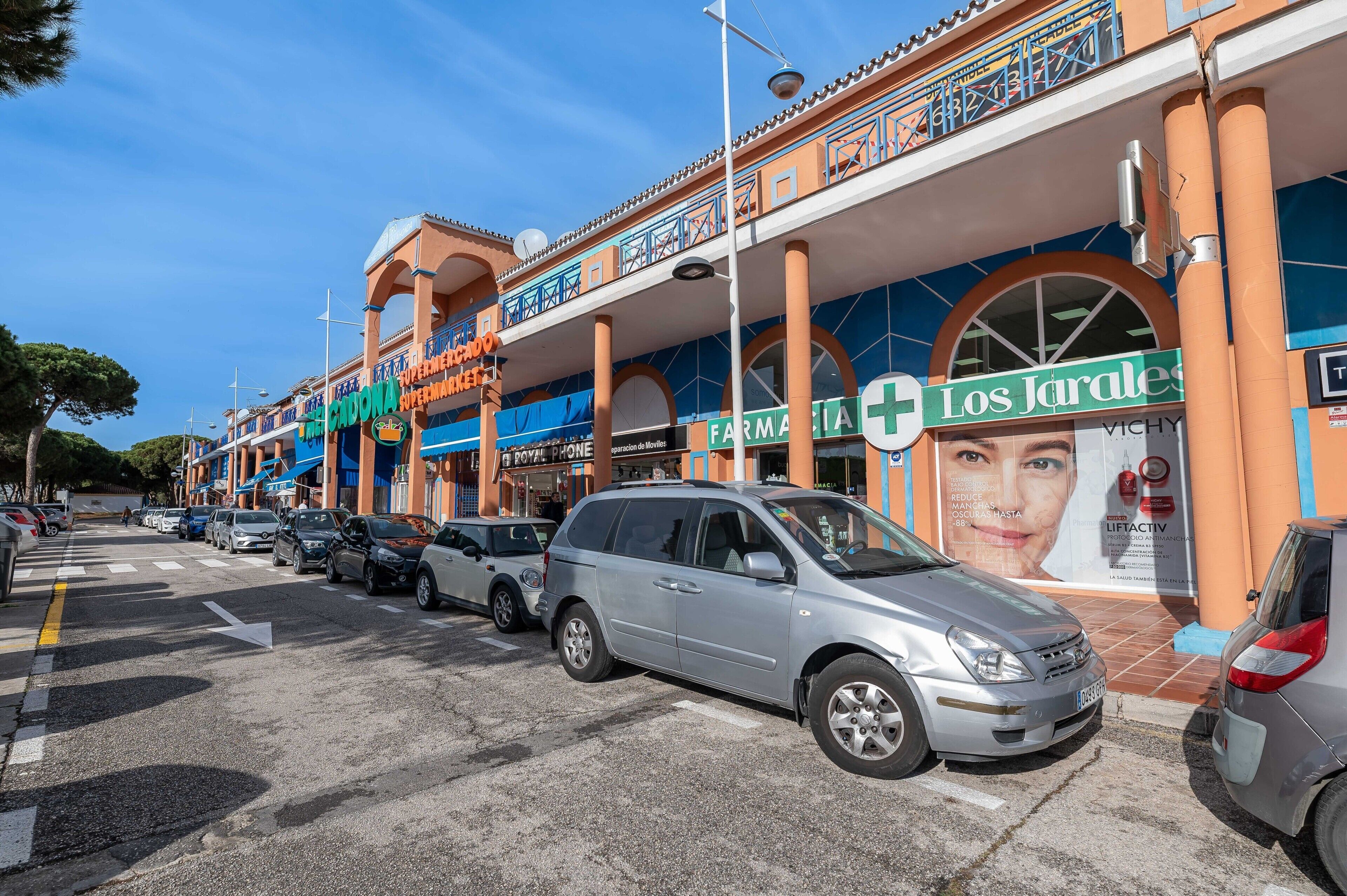 Una calle con coches aparcados y un edificio que alberga una farmacia y un supermercado.