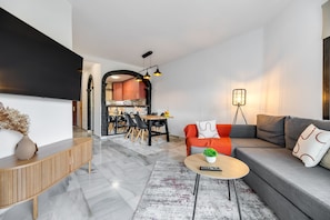 Una sala de estar moderna con sofá, mesa de centro y televisor. Se aprecia un comedor con sillas y una cocina al fondo.