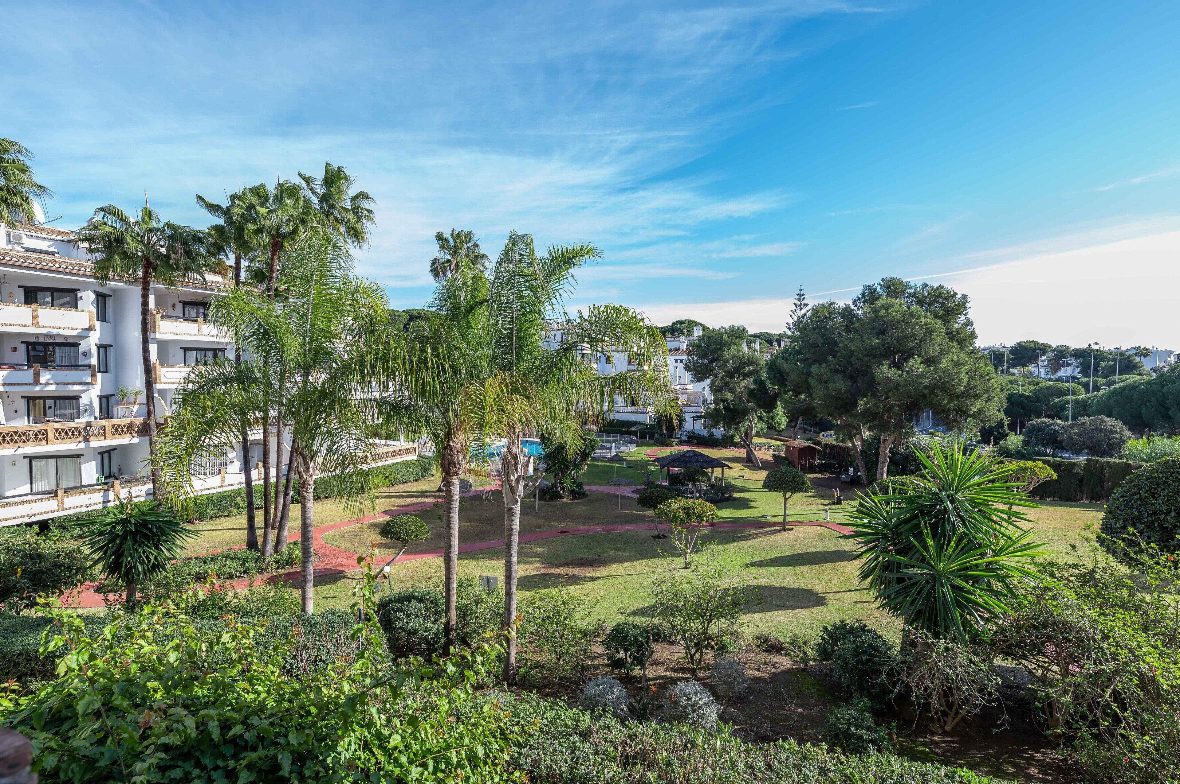 A hotel with multiple floors, a well-maintained garden with palm trees, and a clear blue sky.