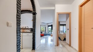 A hallway with a kitchen area, a wooden door, and a view into a living room with a sofa and a window.