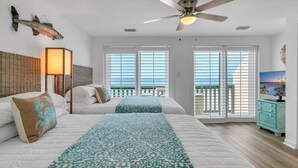 A hotel room with two beds, a ceiling fan, a view of the ocean through large windows, and a wooden floor.