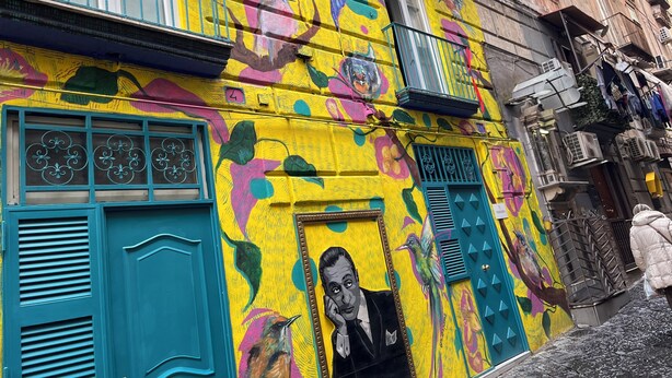Un bâtiment aux couleurs vives, orné de fresques murales éclatantes, une grande fenêtre avec un portrait encadré et un balcon.