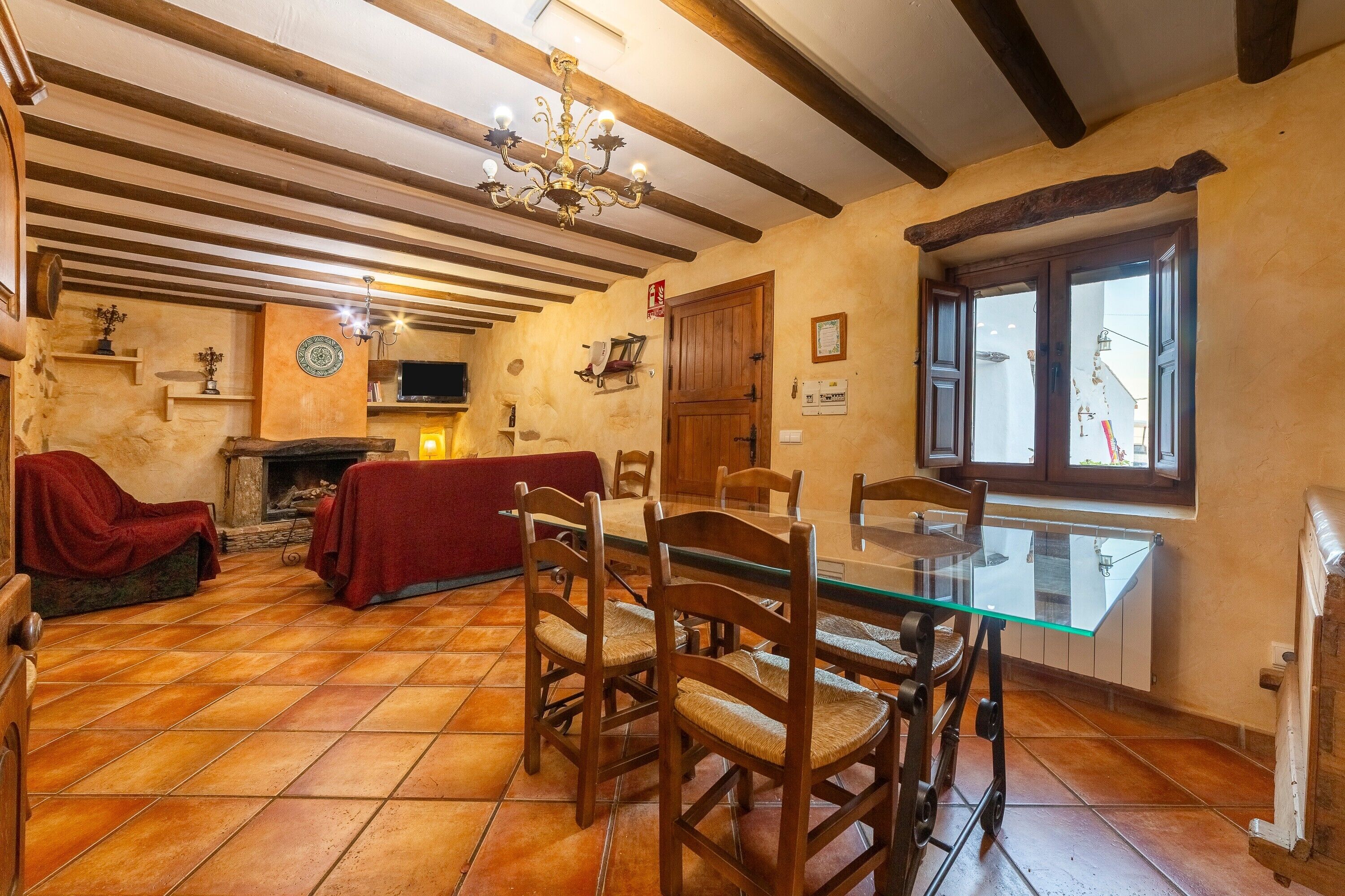 Un comedor con mesa de vidrio, sillas de madera, chimenea y techo de madera con vigas a la vista.