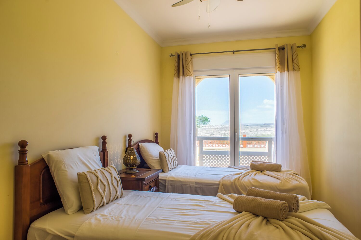 A bedroom with two beds, a nightstand, a lamp, and a view of the outdoors through a large window.