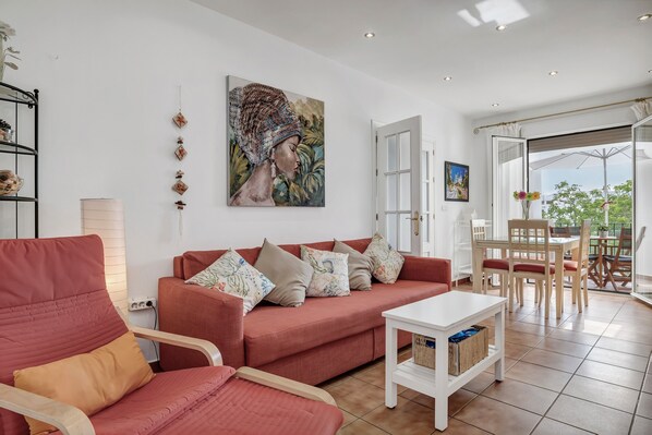 Una sala de estar con un sofá rojo, una mesa blanca y un comedor con sillas de madera y una puerta de vidrio que da al exterior.
