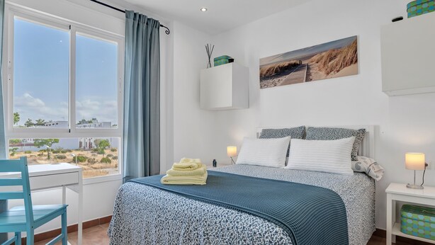 A bedroom with a bed, a desk, a chair, a window with a view, and a nightstand with a lamp.