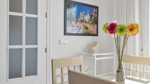 A room with a white door, a framed picture on the wall, a table with chairs, and a vase with flowers.