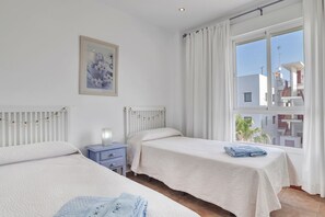 Un dormitorio con dos camas, una mesita de noche, una ventana con cortinas y vistas a edificios.