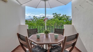 A round table with four chairs and a white umbrella, set on a balcony with a view of trees and buildings.