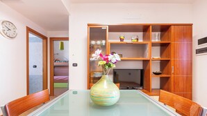 A dining area with a glass-top table, a wooden chair, a vase with flowers, and a wooden cabinet with shelves.