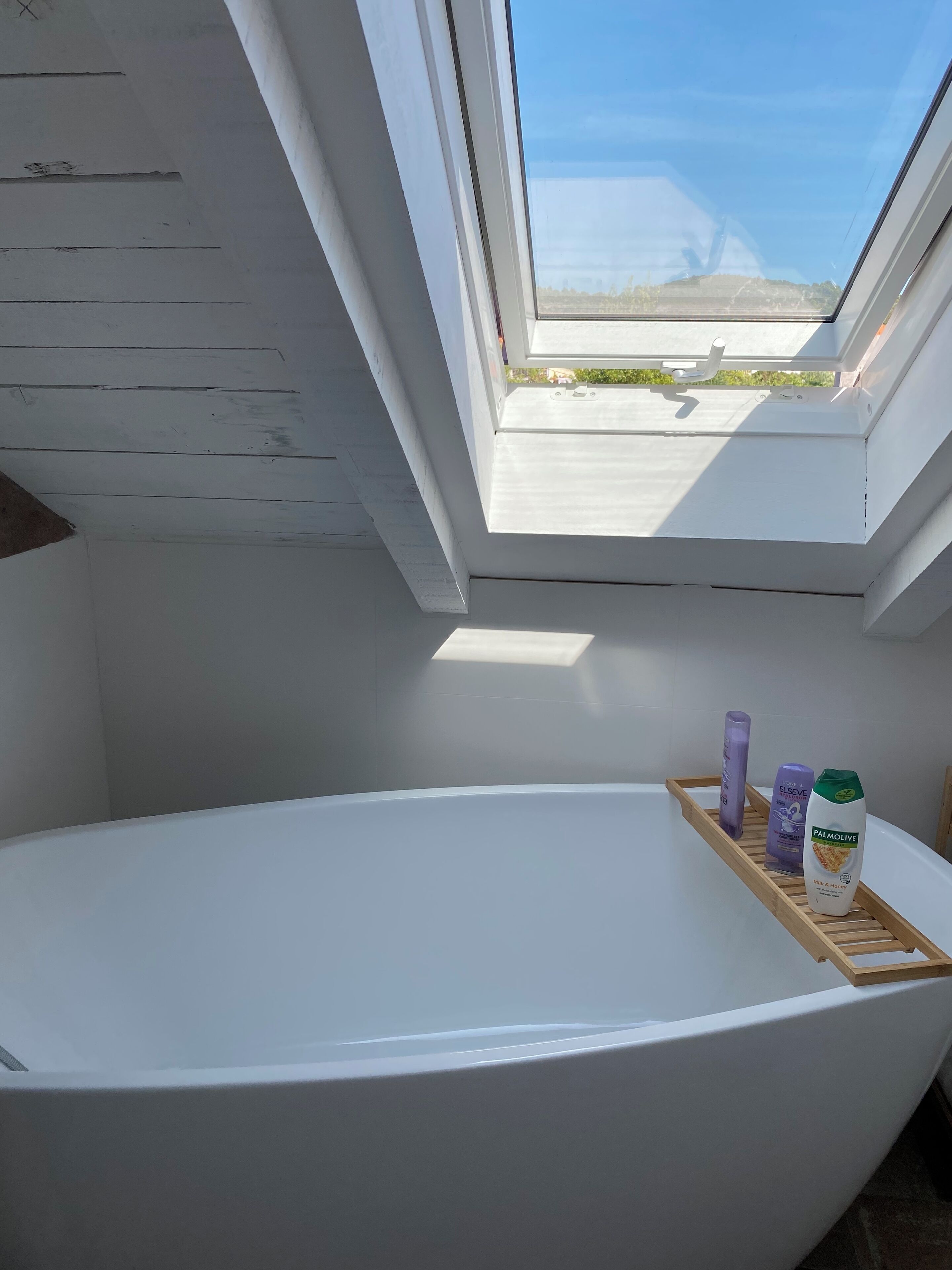 A modern bathroom with a skylight, a freestanding bathtub, and toiletries on a wooden tray.