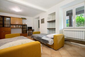 A bedroom with two beds, a wooden cabinet, a radiator, and a window with a view of greenery.