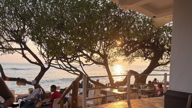 Et udendørs opholdsområde med borde og stole, med udsigt over havet og en solnedgang.