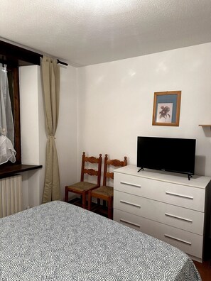Un dormitorio con cama, televisor sobre un aparador, una silla, ventana con cortinas e imagen enmarcada en la pared.