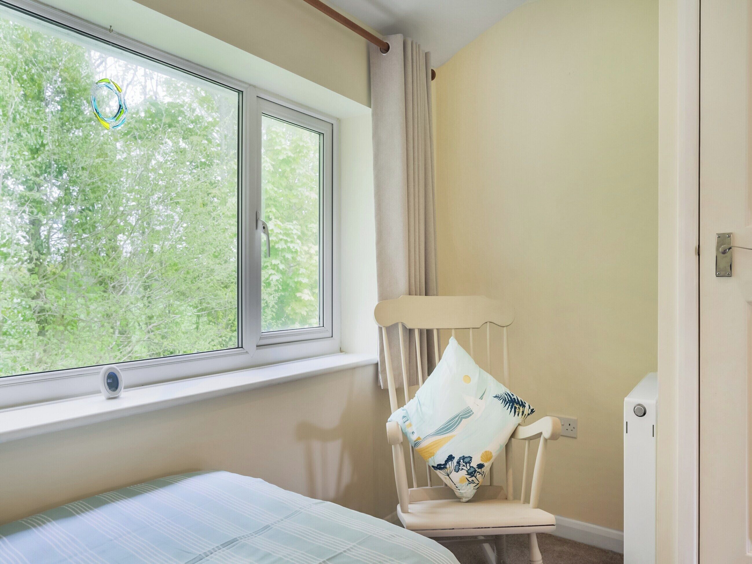 A room with a bed, a window with a view of trees, a chair with a cushion, and a door.