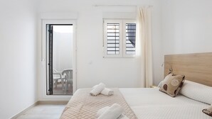 A bedroom with a bed, a window with blinds, a door leading to a balcony, and a wall-mounted light fixture.