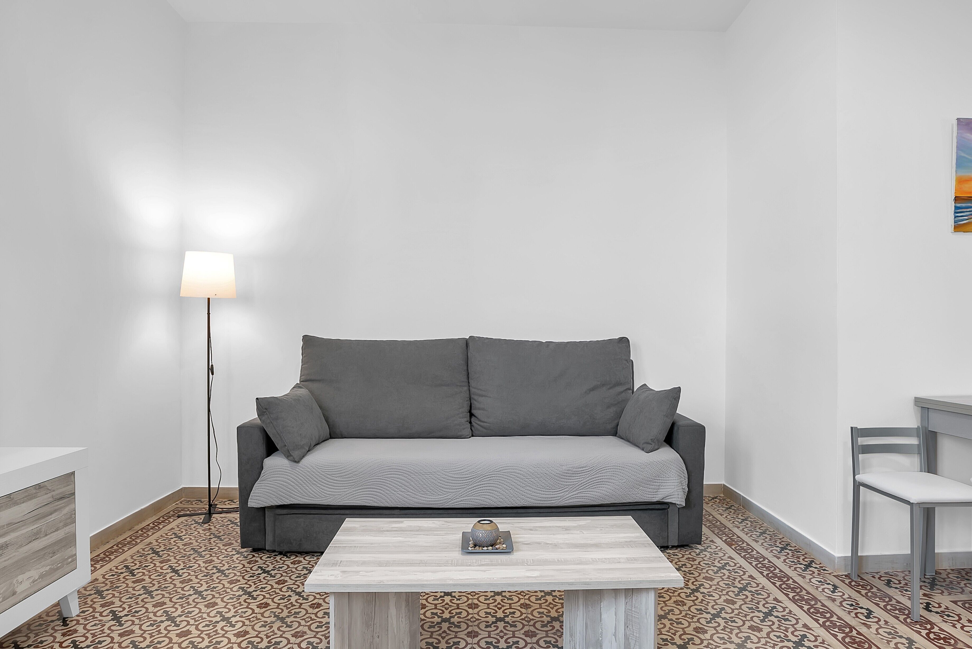 Une pièce avec un canapé gris, une petite table ornée d’un objet décoratif, un lampadaire et un tapis à motifs.