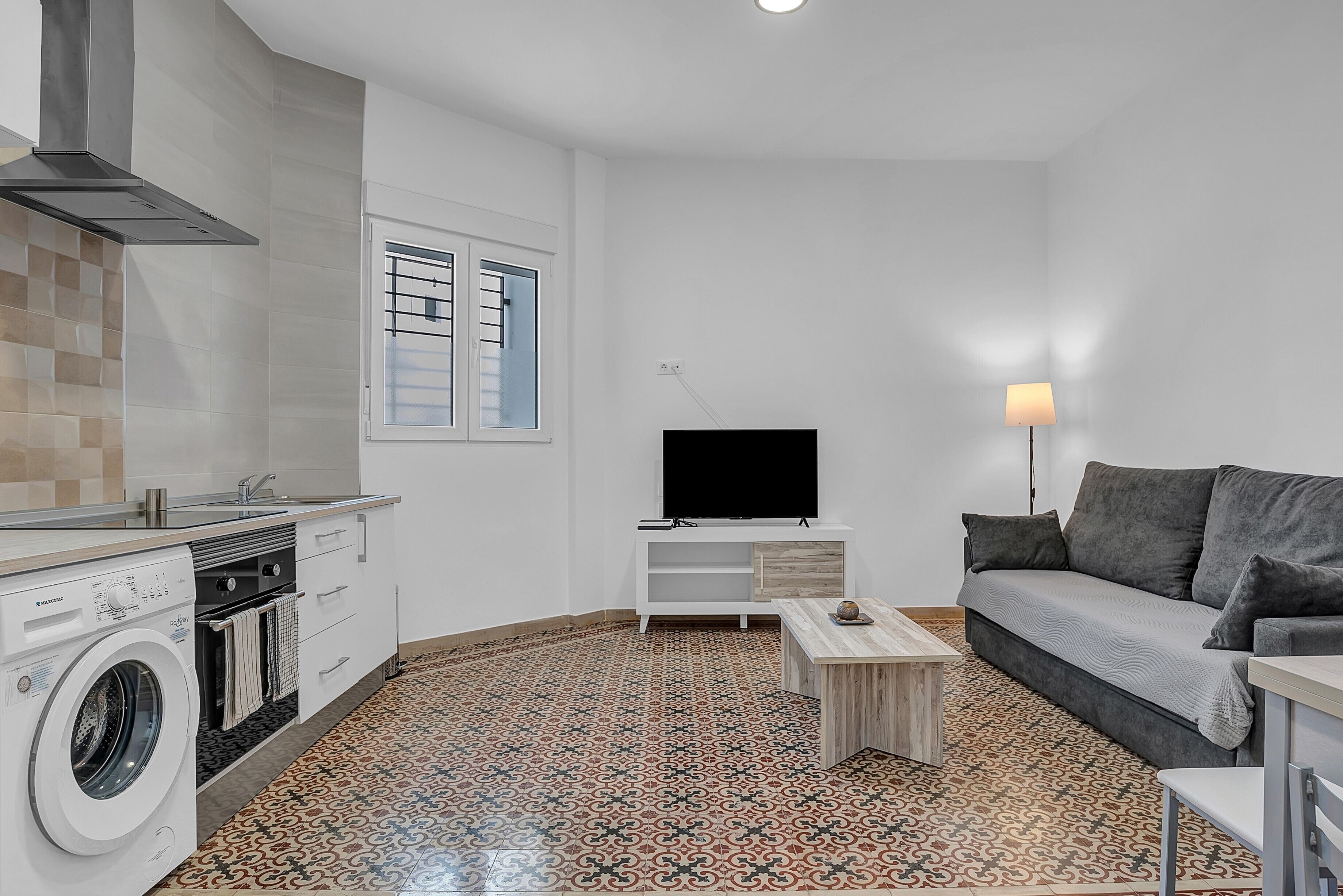Un espace de vie compact comprenant un lave-linge, un téléviseur à écran plat, un canapé et un sol à motifs.