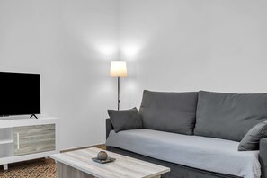 Una sala de estar moderna con un sofá gris, un televisor de pantalla plana sobre un mueble blanco, una lámpara de pie y una mesa de centro con un objeto decorativo.