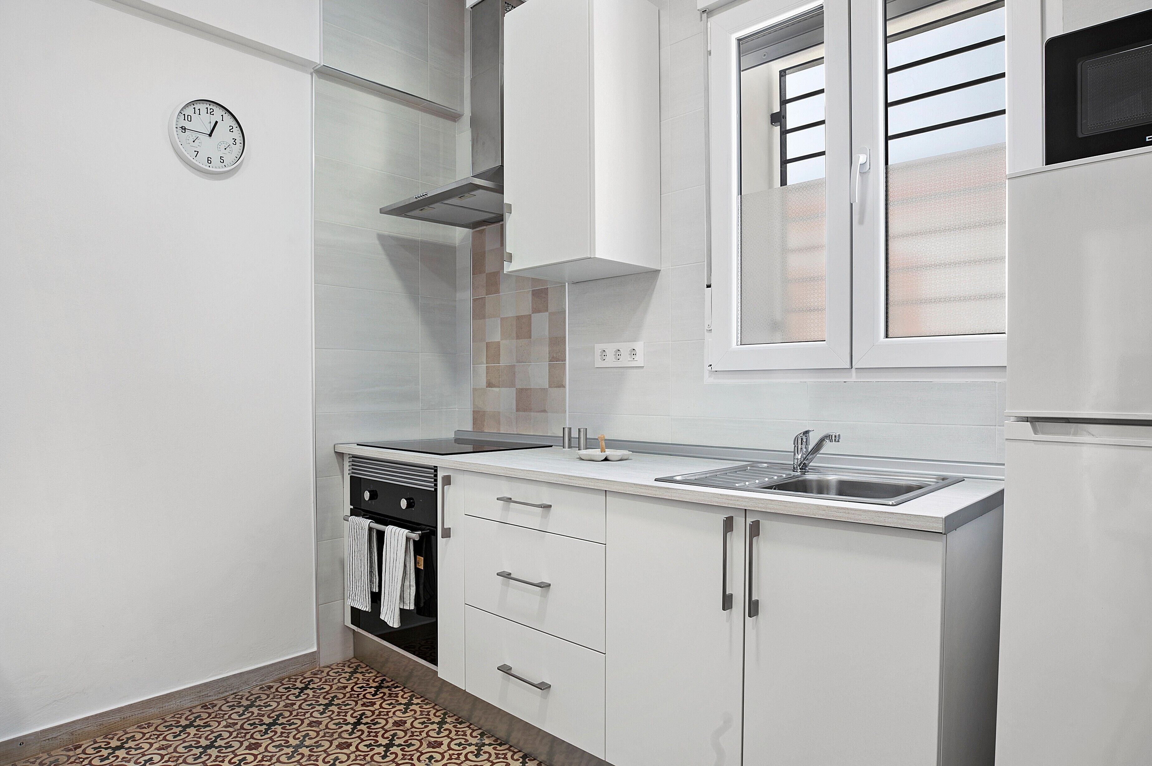 Una cocina con armarios blancos, un lavamanos de acero inoxidable, un reloj y un suelo estampado.