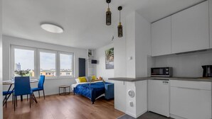 A modern apartment with a dining area, a bed with a blue headboard, a kitchen with white cabinets, and a view of buildings through large windows. - Warsaw Wola Studio - 9th Floor, City View - by Rentujemy (Warszawa)