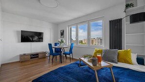 A modern hotel room with a large bed, a dining table with chairs, a TV mounted on the wall, and a city view through the windows. - Warsaw Wola Studio - 9th Floor, City View - by Rentujemy (Warszawa)