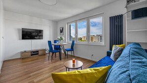 A modern living room with a sofa, a dining table with chairs, a TV mounted on the wall, and large windows offering a city view. - Warsaw Wola Studio - 9th Floor, City View - by Rentujemy (Warszawa)