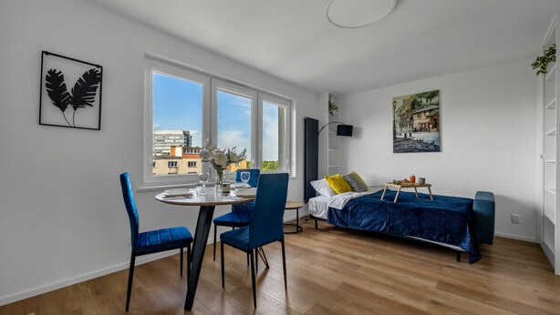A modern apartment with a dining area, a bed with a blue cover, and a city view through large windows. - Warsaw Wola Studio - 9th Floor, City View - by Rentujemy (Warszawa)