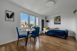 A modern apartment with a dining area, a bed with a blue cover, and a city view through large windows.
