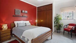 Un dormitorio con cama, armario de madera, mesitas de noche, alfombra roja, planta en maceta y una silla.