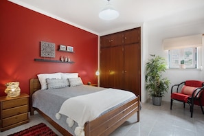 Un dormitorio con cama, armario de madera, mesitas de noche, alfombra roja, planta en maceta y una silla.
