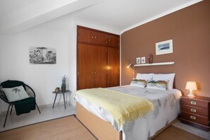 A bedroom with a bed, a chair, a small table, a wardrobe, and a bedside table with a lamp. - Apartment 'Porto Dinheiro Beach 1' with Mountain View, Shared Pool and Wi-Fi (Ribamar-Lourinhã)
