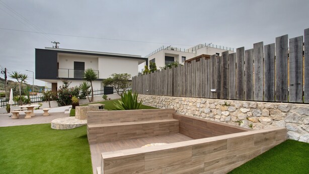 Un área de estar al aire libre y de diseño moderno, con una pared de piedra y terraza de madera, rodeada por un césped cuidado y una valla de madera.