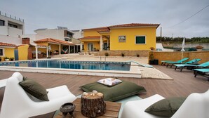 Zona de piscina con sillones de descanso, una mesa de madera y un edificio amarillo al fondo.