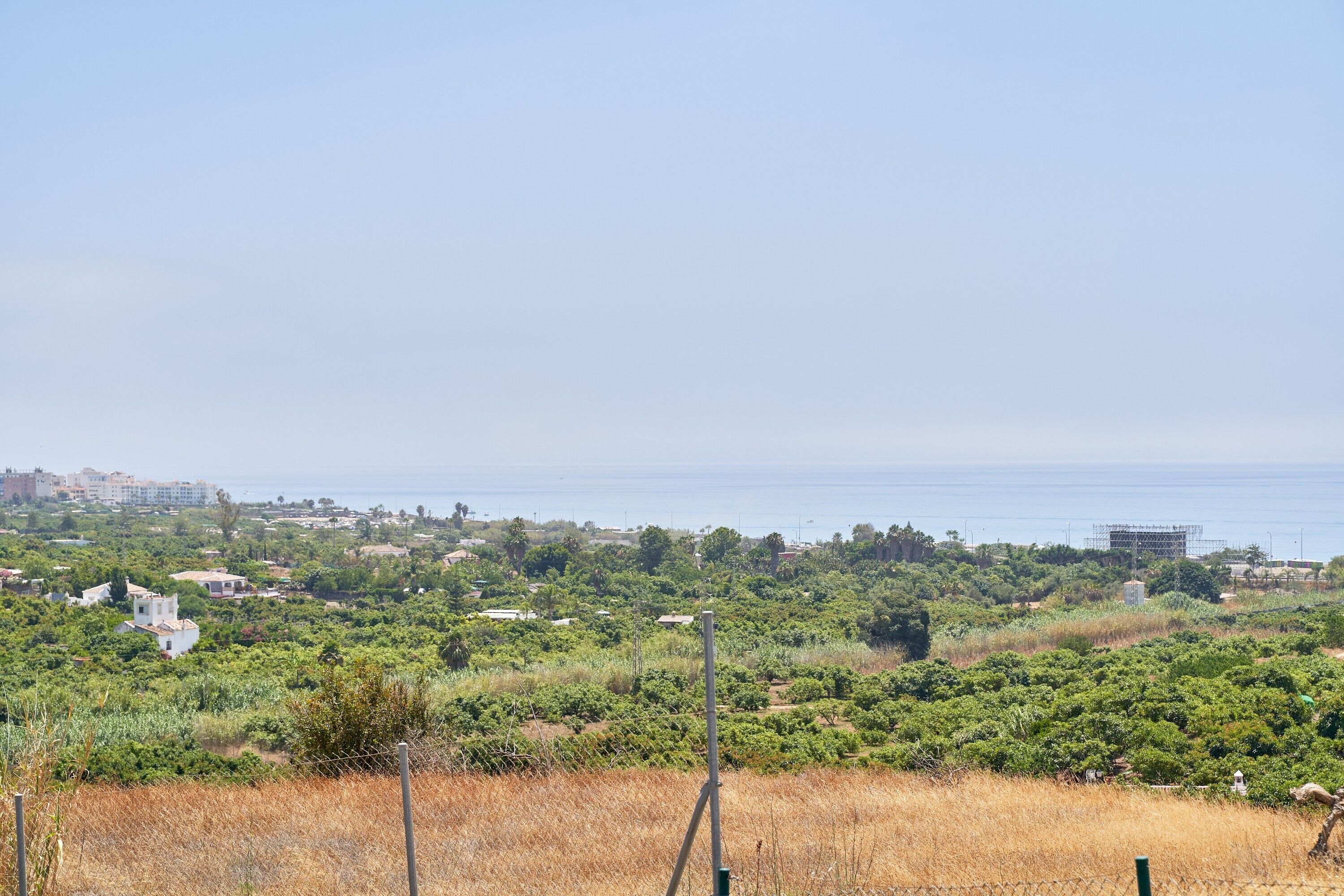 A coastal landscape with a view of the sea, a fence, and scattered buildings.