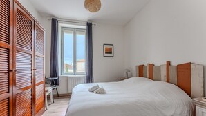 A bedroom with a large bed, wooden shutters, a window with curtains, a chair, and a small table with a lamp.