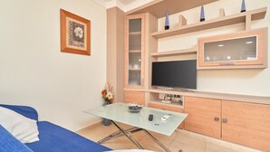 Un salon moderne avec une table basse en verre, un canapé bleu, un téléviseur et un meuble contenant des objets décoratifs.
