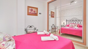 Une chambre avec un grand miroir, un lit avec une literie rose, une chaise et une petite table.