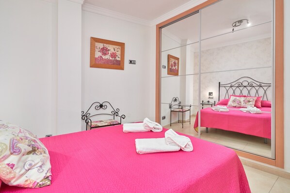 Une chambre avec un grand miroir, un lit avec une literie rose, une chaise et une petite table.