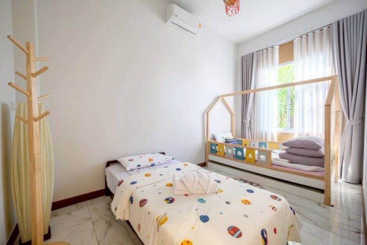 A bedroom with a bed, a wooden ladder, a window with curtains, and a small table with pillows.