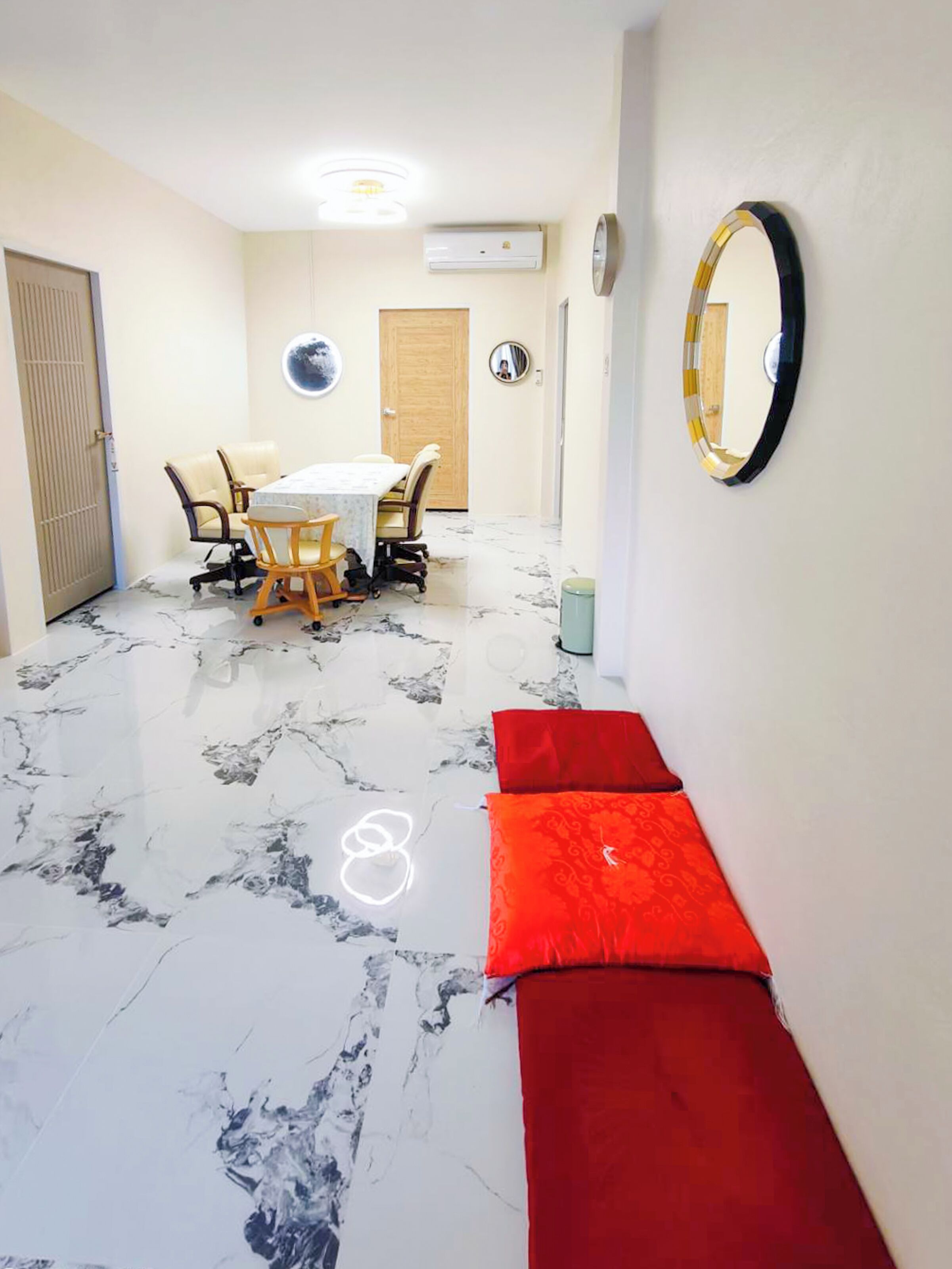 Una habitación con piso de mármol, un banco rojo, un espejo redondo y una zona de comedor con sillas y una mesa.