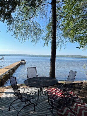 Un patio junto al lago con sillas y una mesa de metal, un muelle de madera que se adentra en el agua, y una vista despejada del lago y colinas distantes.