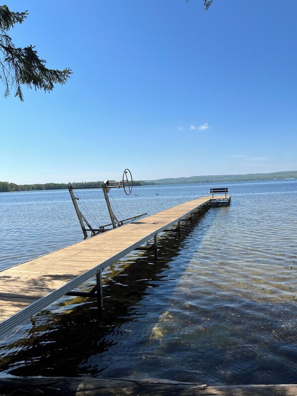 Un muelle de madera que se adentra en un lago tranquilo, con cielo despejado y un barco a lo lejos en el agua.